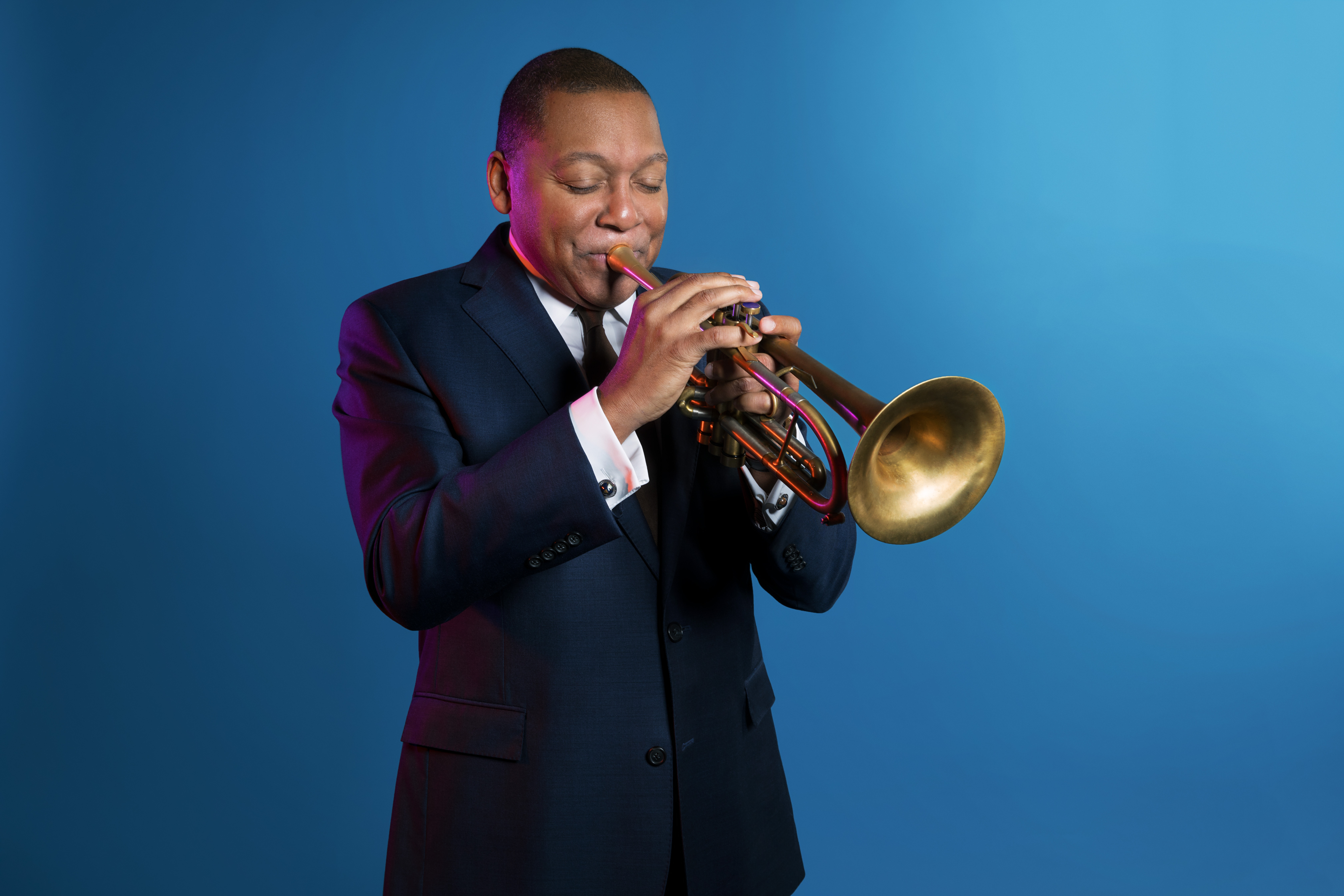 Wynton Marsalis, wearing a suit and playing a trumpet. Blue studio background 