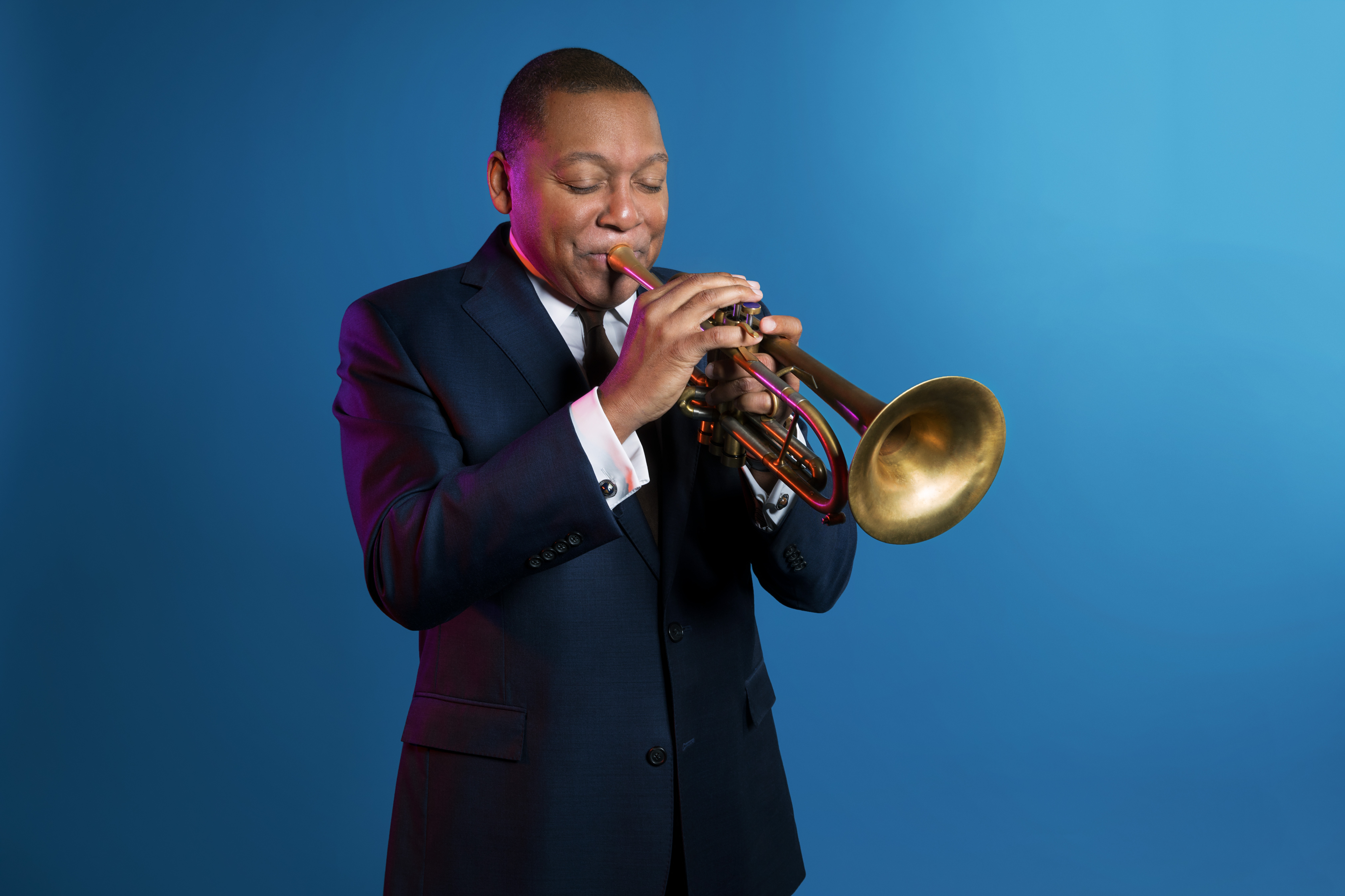 Wynton Marsalis, wearing a suit and playing a trumpet. Blue studio background