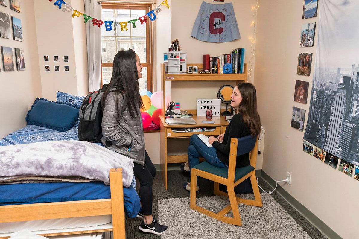 Two students chat in a student residence, one seated at a desk, the other leaning on the bed