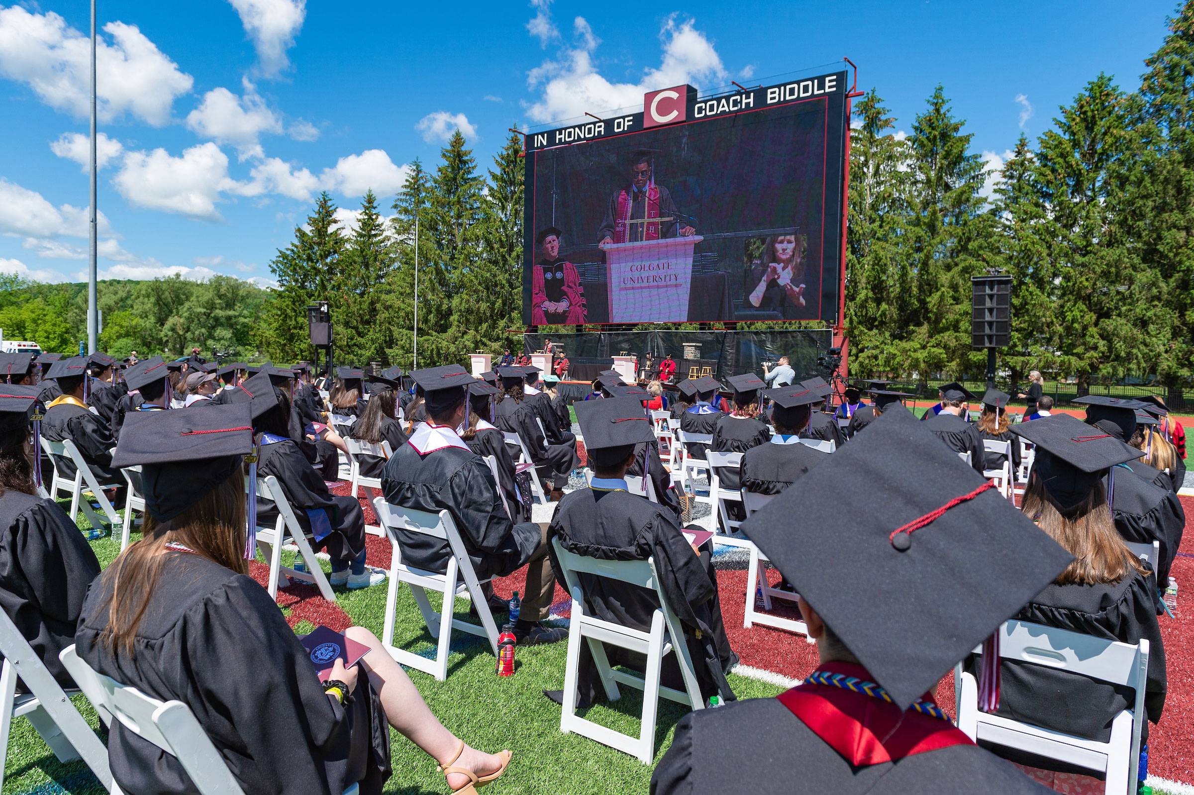graduates in caps and gowns sitting in white folding chairs on the football field, ceremony on stadium screen in background