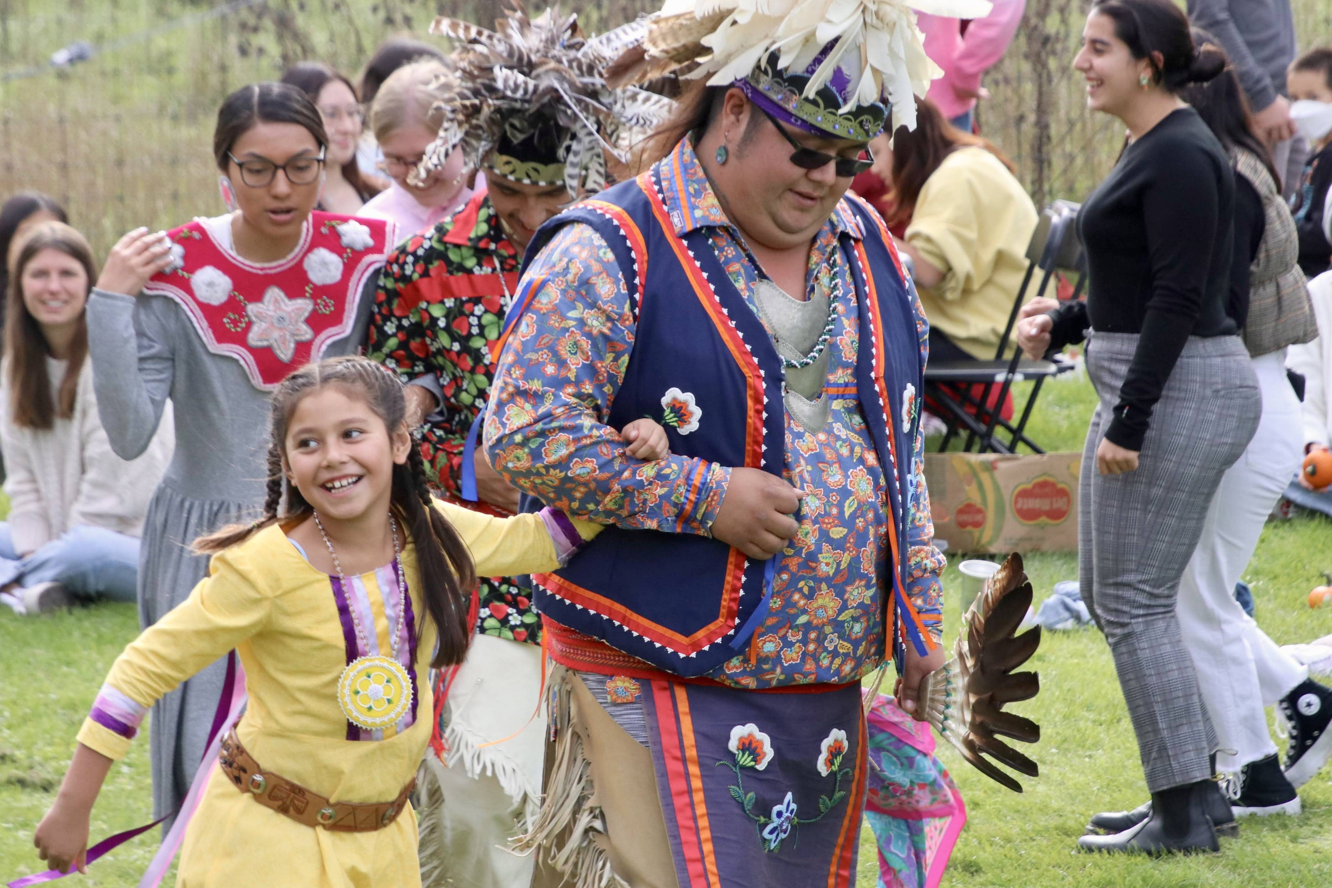 A girl in a yellow dress links arms with one of the Haudenosaunee dancers at the Fall Festival.