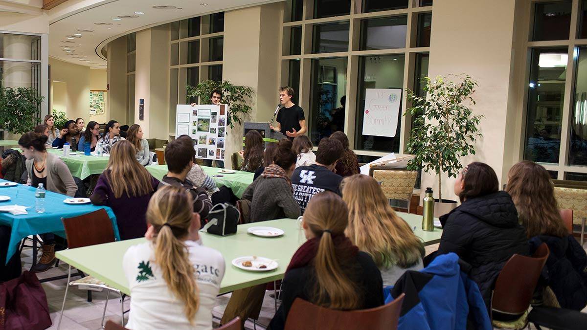 Students and community members seated at tables listening to a speaker at the front of the audience.