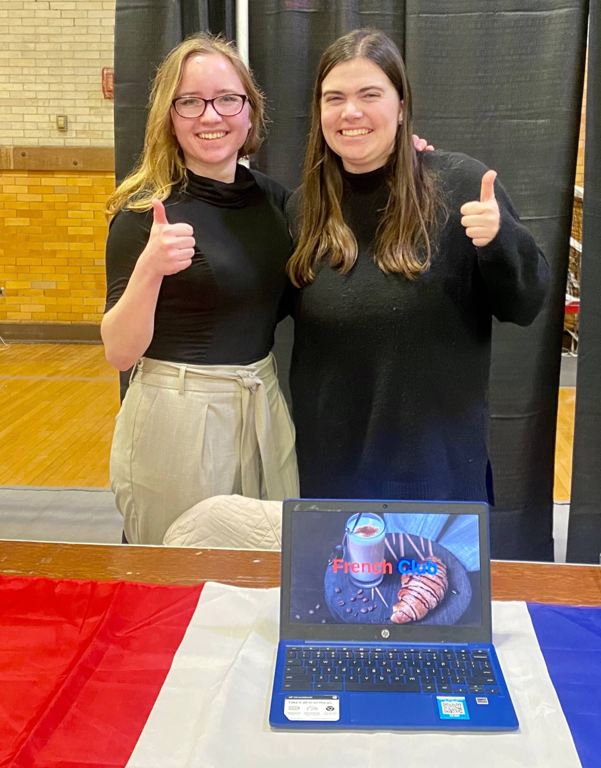 Two Colgate students pose with thumbs up signs in front of a laptop, sitting on table decorated with the colors of France's flag.