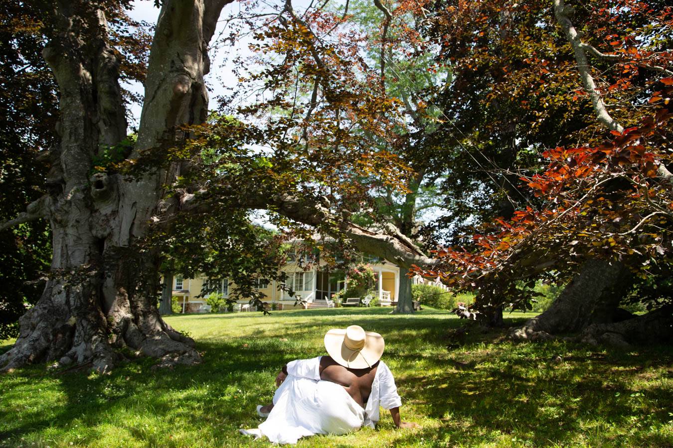 Picture of woman in white dress and hat from the back sitting in woods looking at a yellow house