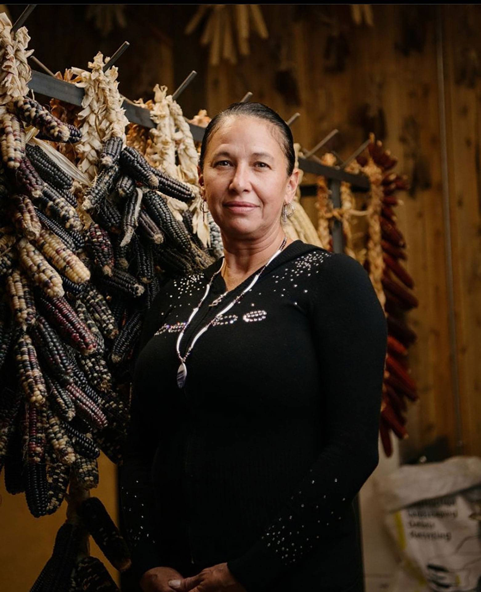 Angela Ferguson poses in front of a rack holding multicolored dried ears of corn strung together