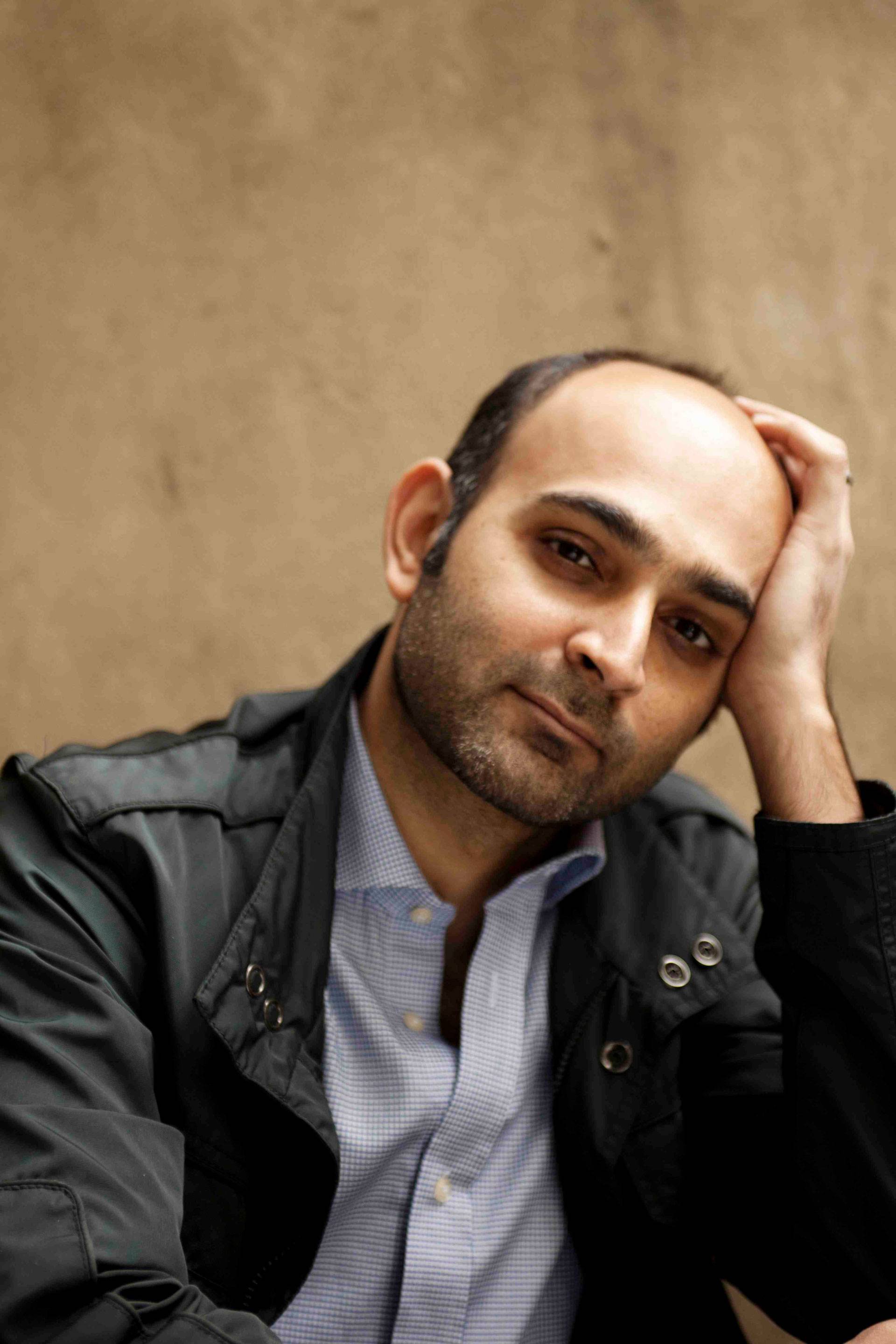 Portrait of Mohsin Hamid leaning his head on one hand, wearing a black jacket and blue-and-white checked shirt