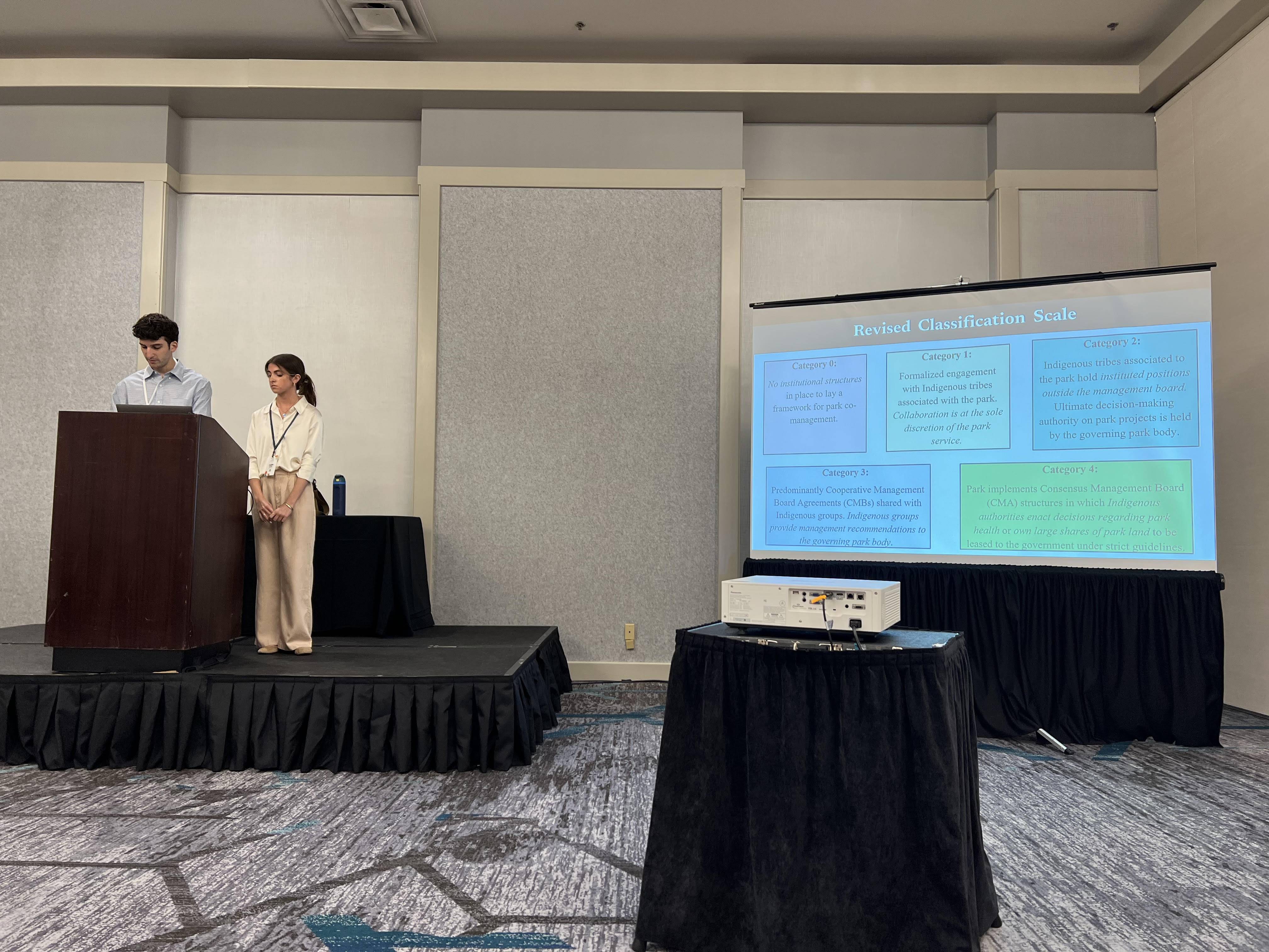 Photo of two students standing at a podium with a screen on their right.
