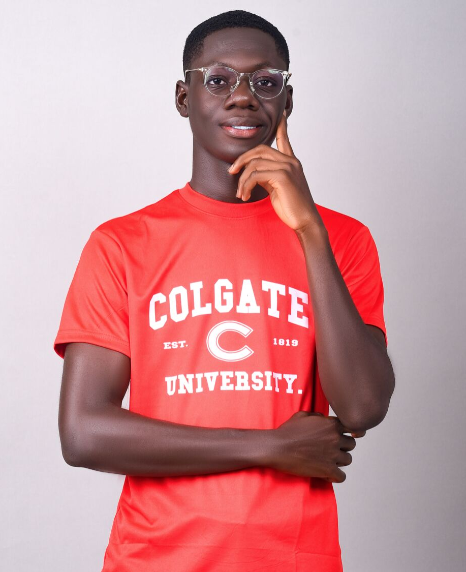 An upper torso photo of Bob Aboh wearing a red Colgate shirt, his right arm across his stomach, and his left arm at his chin, as if thinking.