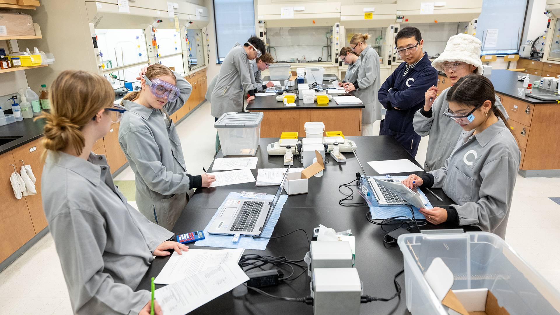 Students wearing protective eye gear and lab coats gather around a table in a science lab classroom