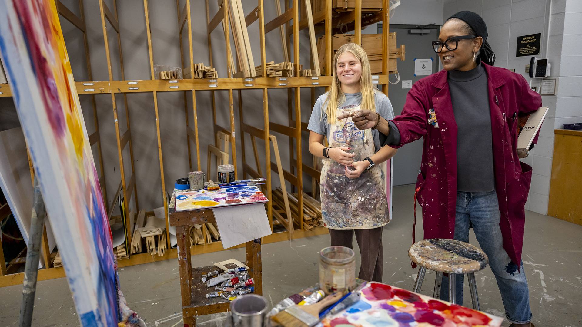An art professor and student smile while looking at a canvas in an art studio.