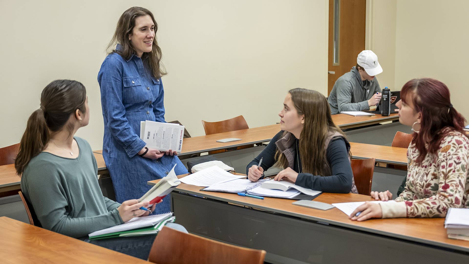A smiling professor leans on a desk and speaks with students sitting at desks.