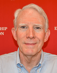 A headshot of a man with short, light-colored hair wearing a blue and white striped button-down shirt. He is looking directly at the camera against a bright red background that features partial white text.