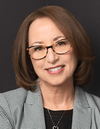 A headshot of a smiling woman with shoulder-length brown hair and glasses with tortoiseshell frames. She is wearing a grey patterned blazer over a black top and a delicate necklace, posed against a solid dark background.