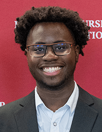 A headshot of a smiling man with dark, textured hair and glasses with thin, orange-accented frames. He is wearing a white button-down shirt under a dark blazer, set against a red background with partial white lettering.
