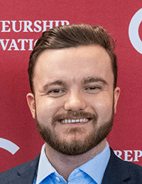 A headshot of a smiling man with brown hair and a neatly trimmed beard, wearing a blue checkered shirt and a dark blazer. He is positioned in front of a red backdrop featuring white text related to entrepreneurship and innovation.