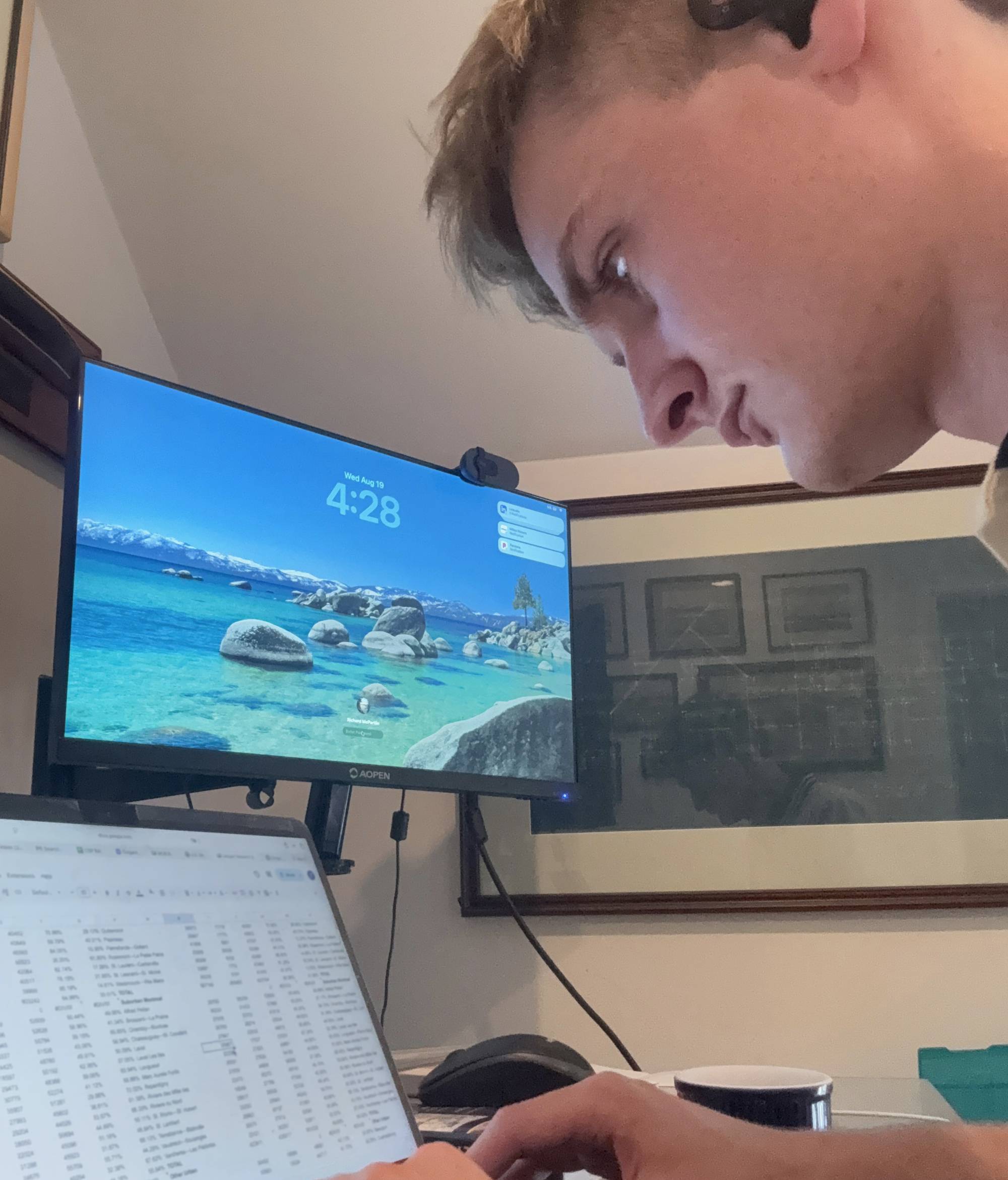 A young man is captured in a side-profile view as he works at a desk, looking down toward a open laptop displaying a spreadsheet. Positioned behind the laptop is an external monitor showing a desktop wallpaper with a scenic beach and mountains.
