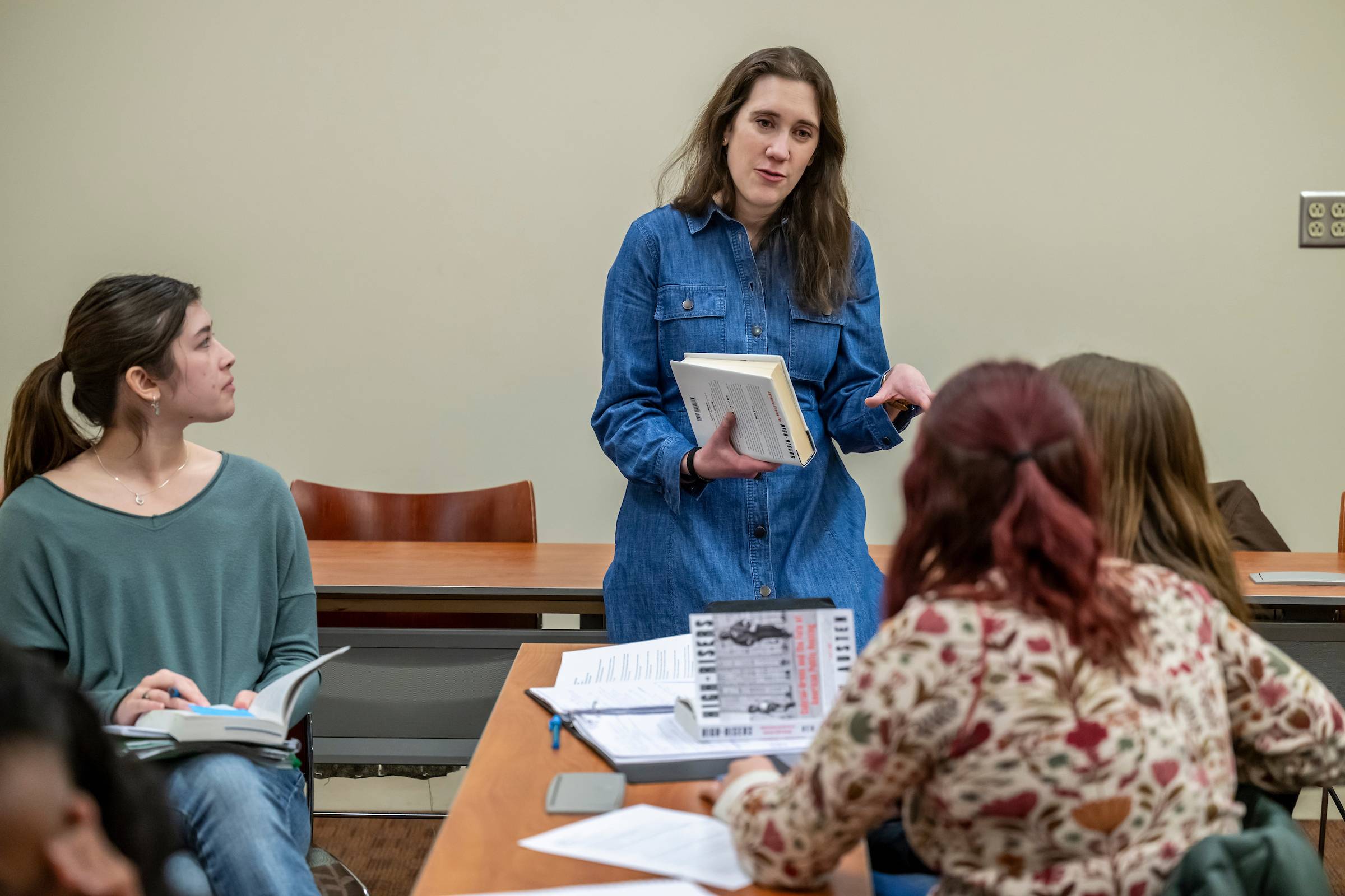A geography professor stands and visits with students sitting at desks in a classroom.