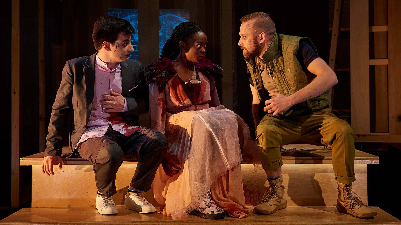 Three actors in costume converse in character while seated on stage