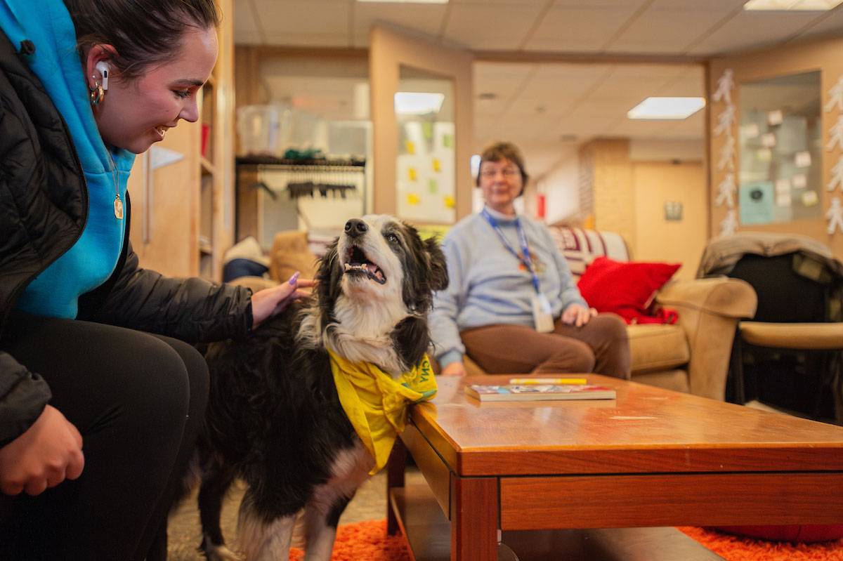 A student, sitting in a chair, pets a black and white dog wearing a yellow kerchief. A woman pet therapy dog handler watches in the background.