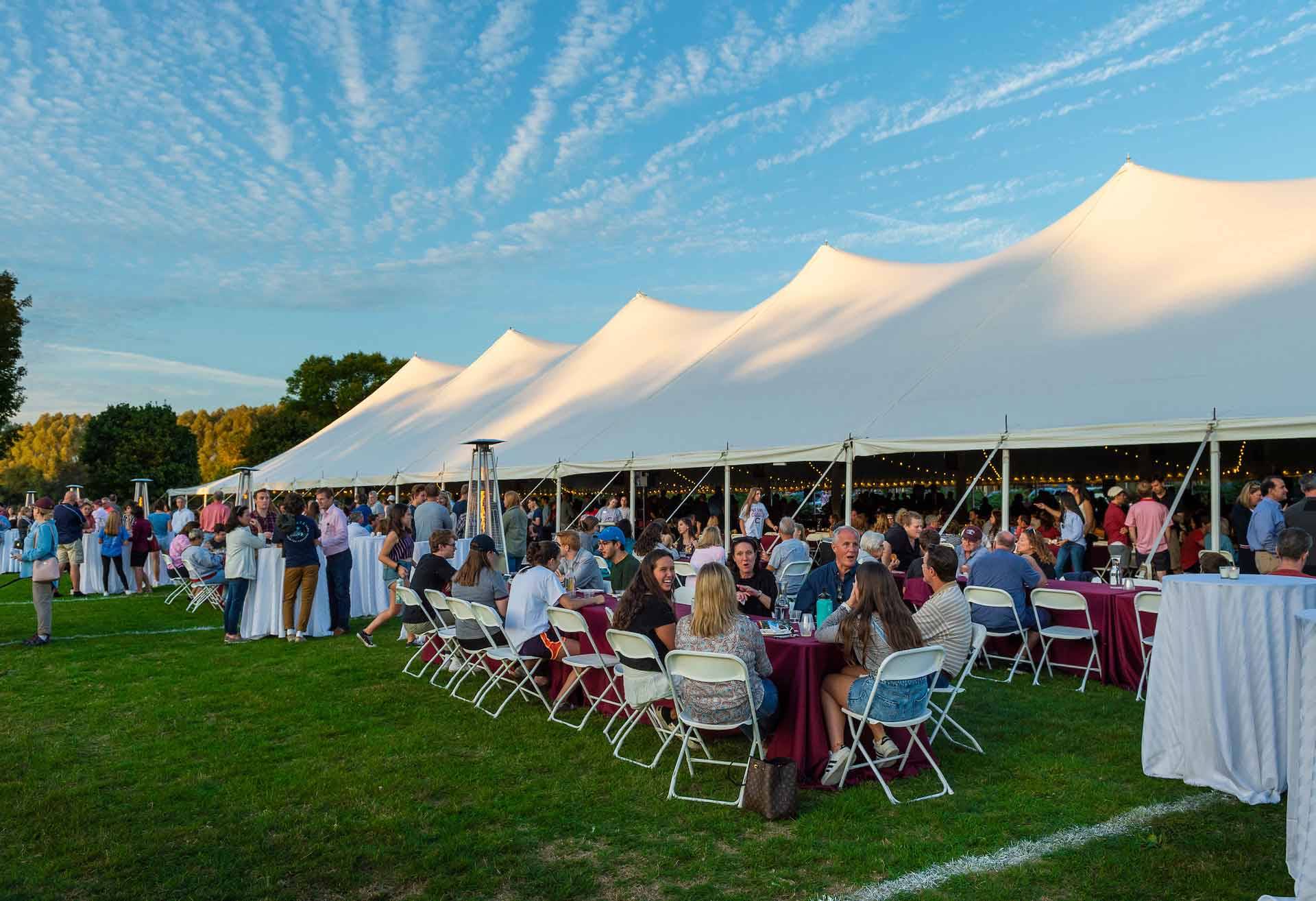Families seated at tables in front of white tent under a blue sky