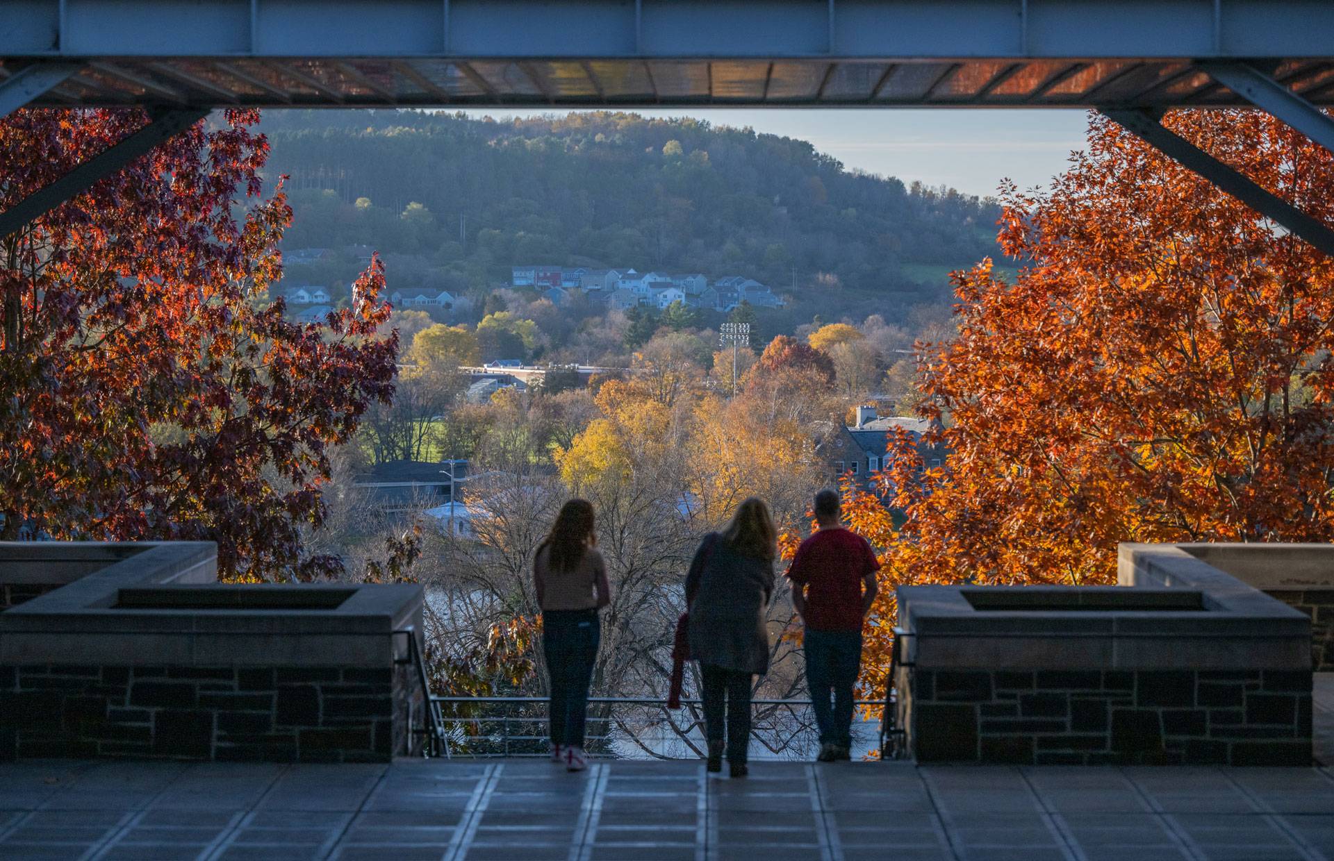 Three people at the edge of the patio under the bridge at Persson Hall, overlooking the lower campus and village with the hills in the distance. Trees in fall color are on either side of them.