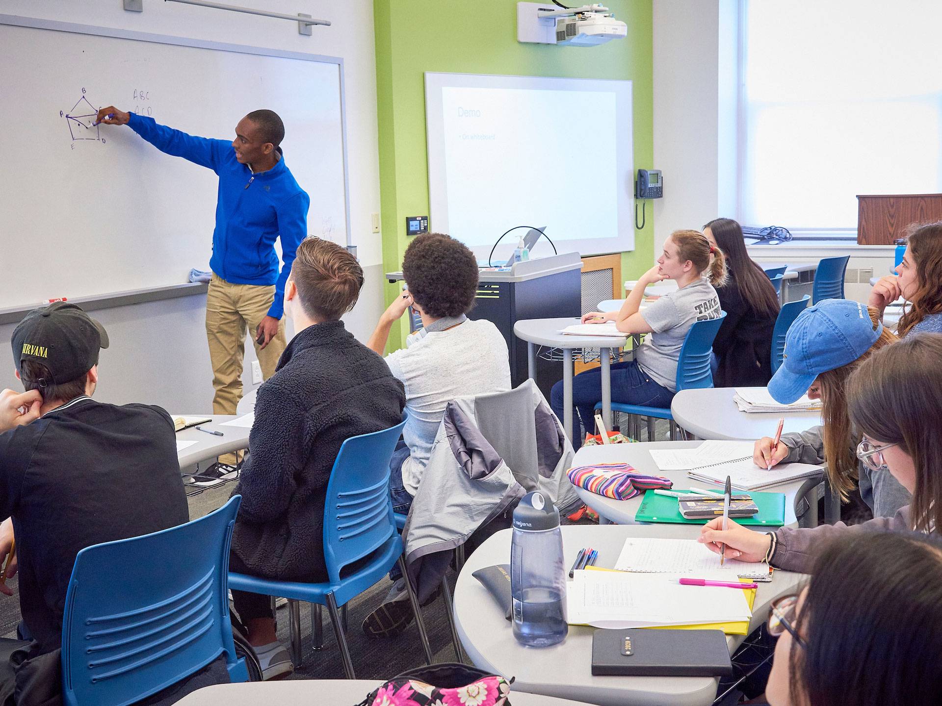 An African American student in a blue zip jacket and chinos draws a diagram on a white board in front of his classmates.