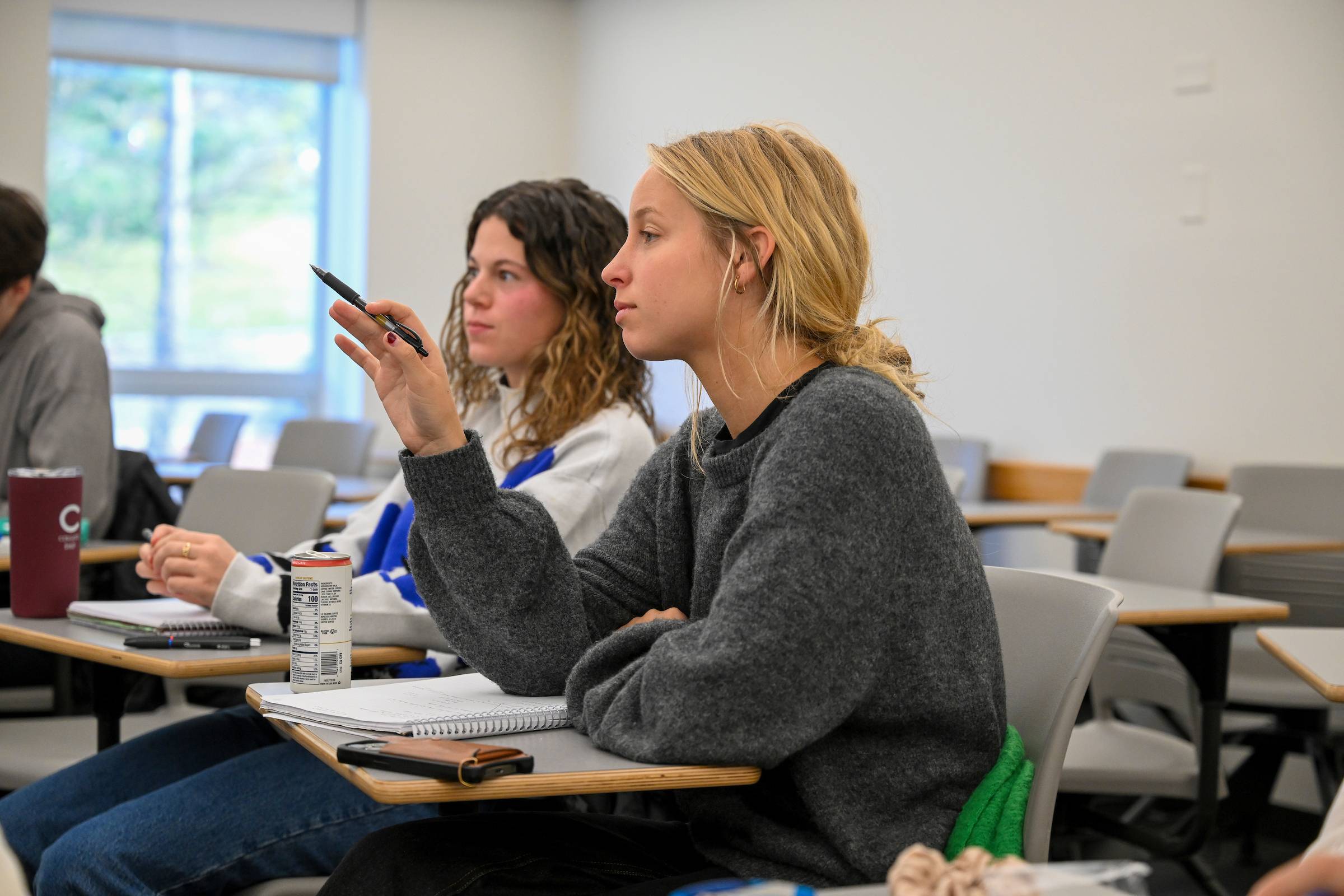 Two students sit at desks in a classroom. One student raises their hand.
