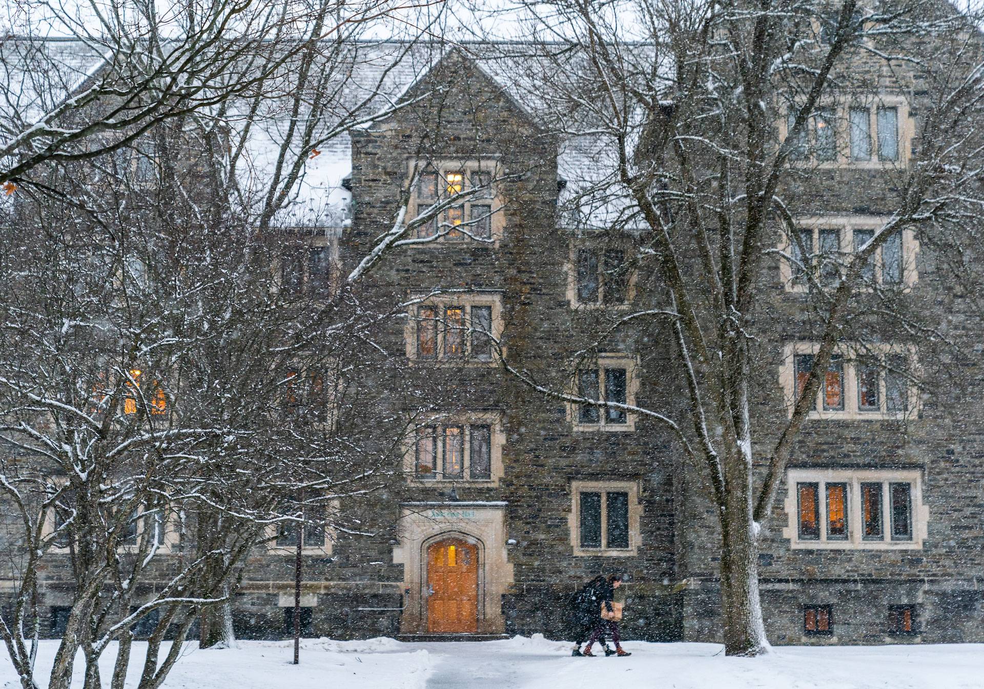 Two students, one carrying a cardboard box, walk during a snowfall in front of Andrews Hall, a stone building with five stories and multiple roof peaks