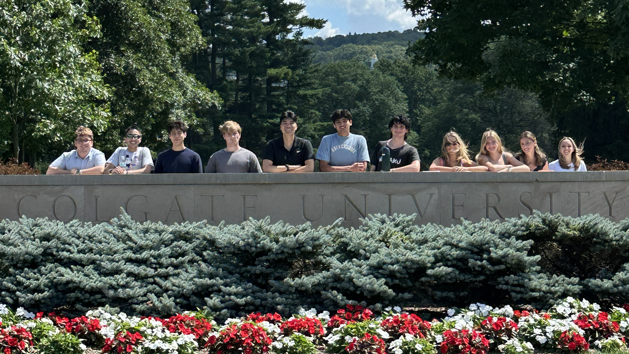 Eleven young adults pose in a row, leaning over a large stone wall engraved with the words "COLGATE UNIVERSITY." A bed of red and white flowers and evergreen shrubs borders the base of the sign, set against a backdrop of dense green trees and a forested hill.