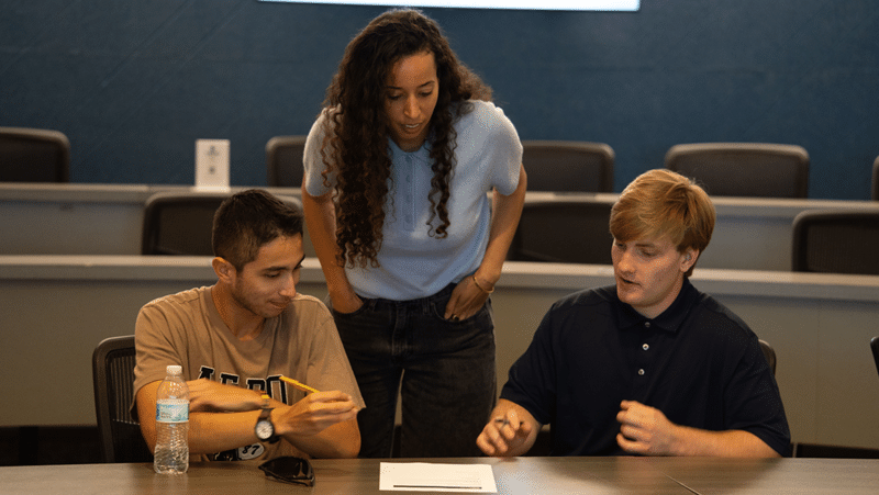 A woman with long, curly dark hair stands leaning over a table in a classroom between two seated young men, all focusing on a document in front of them. The blonde man on the right gestures while speaking, while the man on the left holds a pen near a plastic water bottle on the desk.