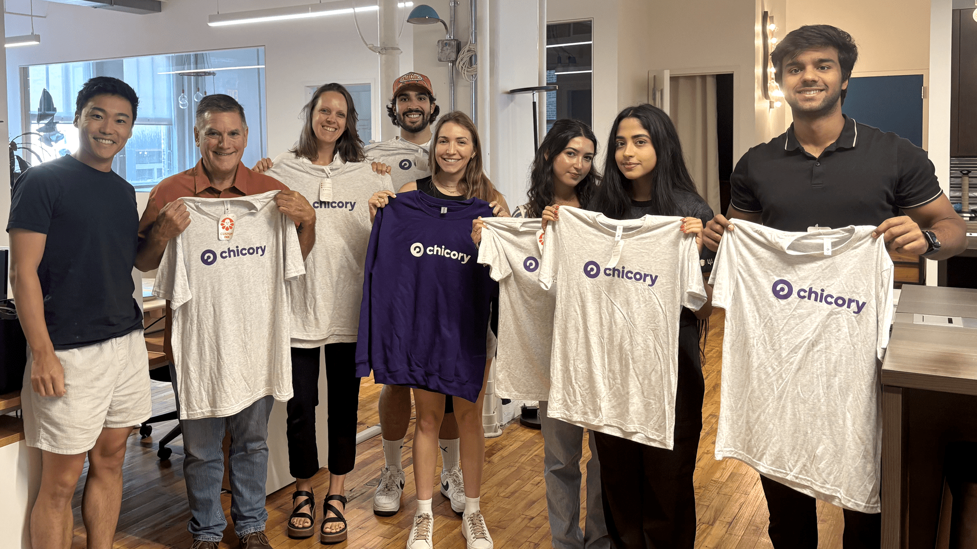 A group of eight smiling individuals stands in a bright, modern office holding up white and purple apparel featuring the "Chicory" logo. The team is diverse and posed in a row across a light wood floor.