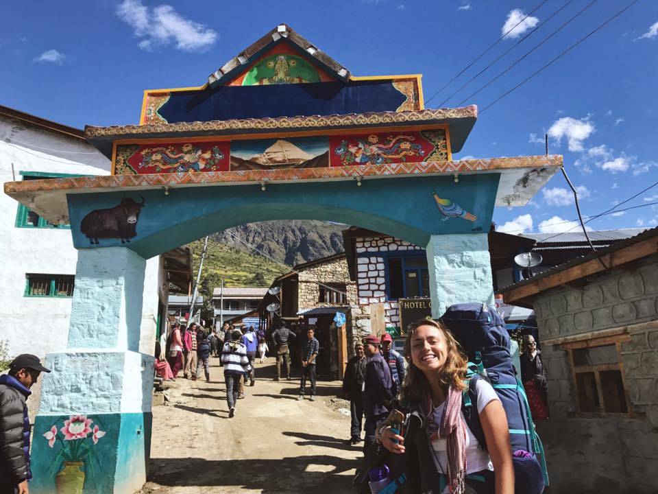 Jenny stands in front of an arch in the Nepalese mountains, wearing a backpack and smiling