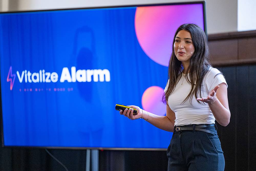 A student entrepreneur presents on stage in front of a large screen displaying the words “Vitalize Alarm,” holding a clicker and gesturing as she speaks. 