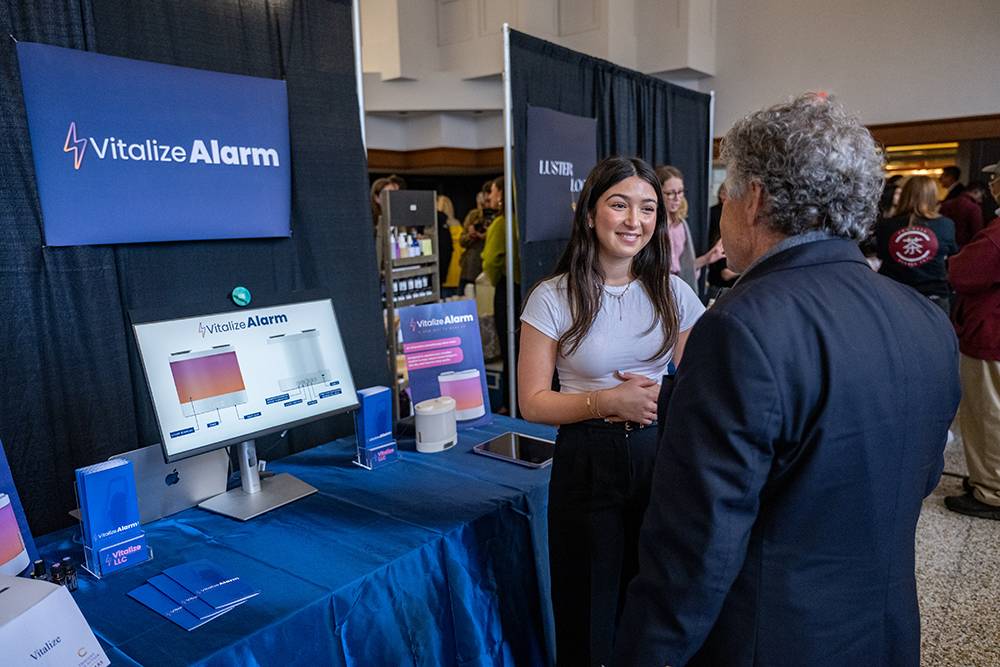 An entrepreneurial student stands beside a display table and speaks with another attendee in front of a booth featuring a monitor, brochures, and a large “Vitalize Alarm” sign.
