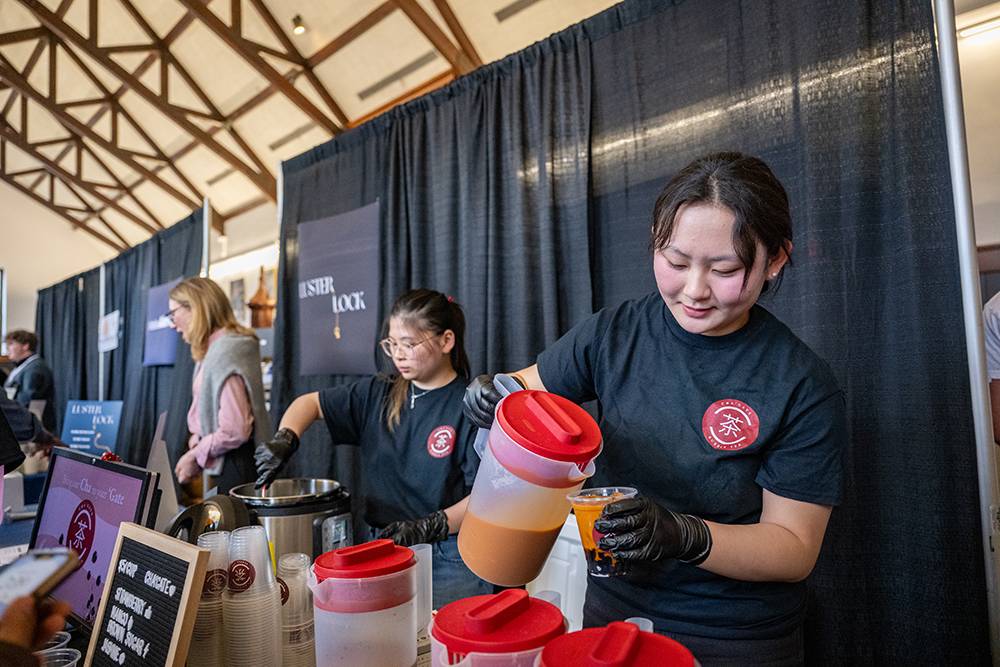 A student wearing a black Cha’Gate T-shirt pours bubble tea from a large pitcher into a plastic cup as another team member works behind the table at the 2025 Entrepreneur Showcase.