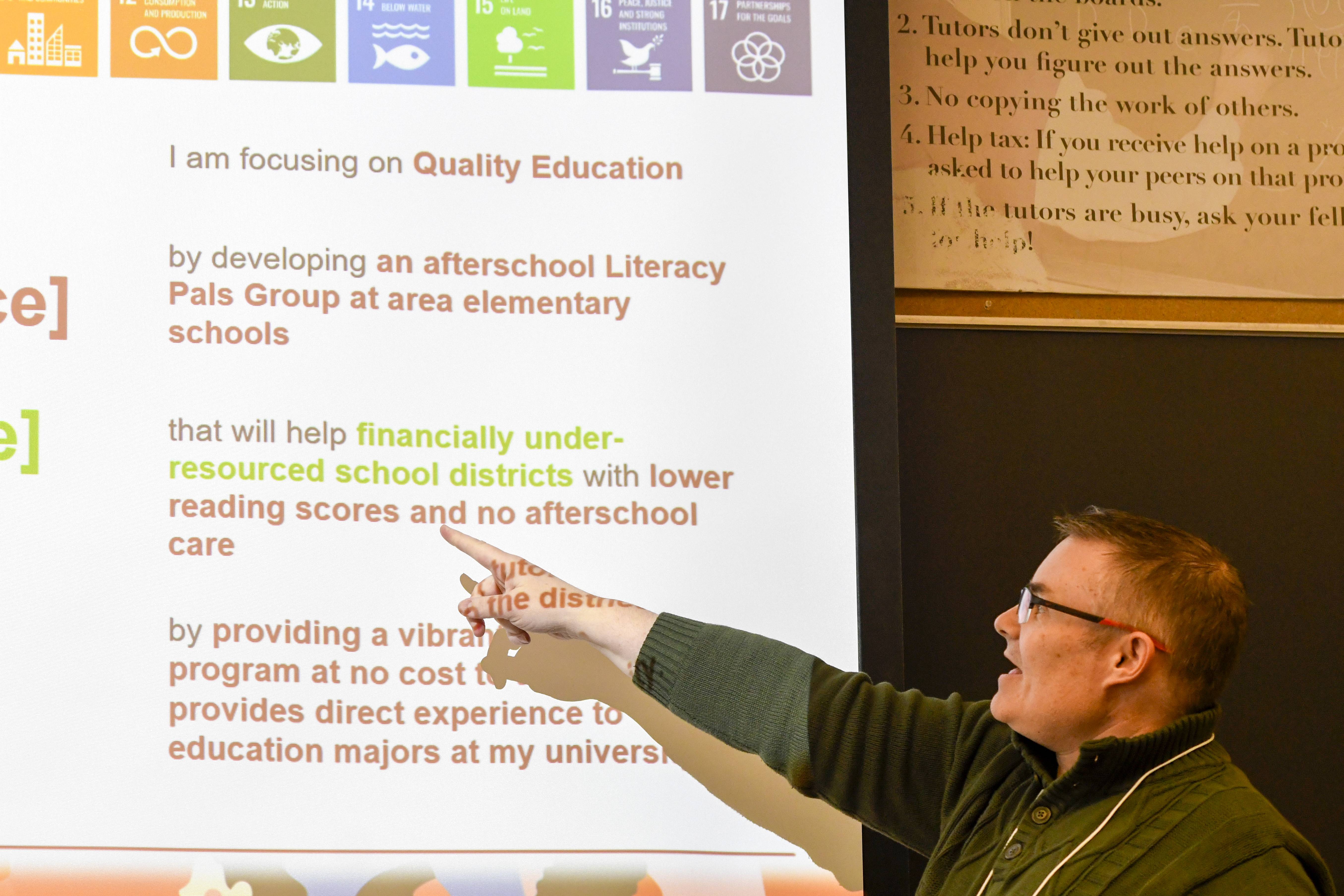 A presenter gestures toward a projected slide about “Quality Education,” describing an afterschool literacy program designed to support under-resourced elementary school districts. 
