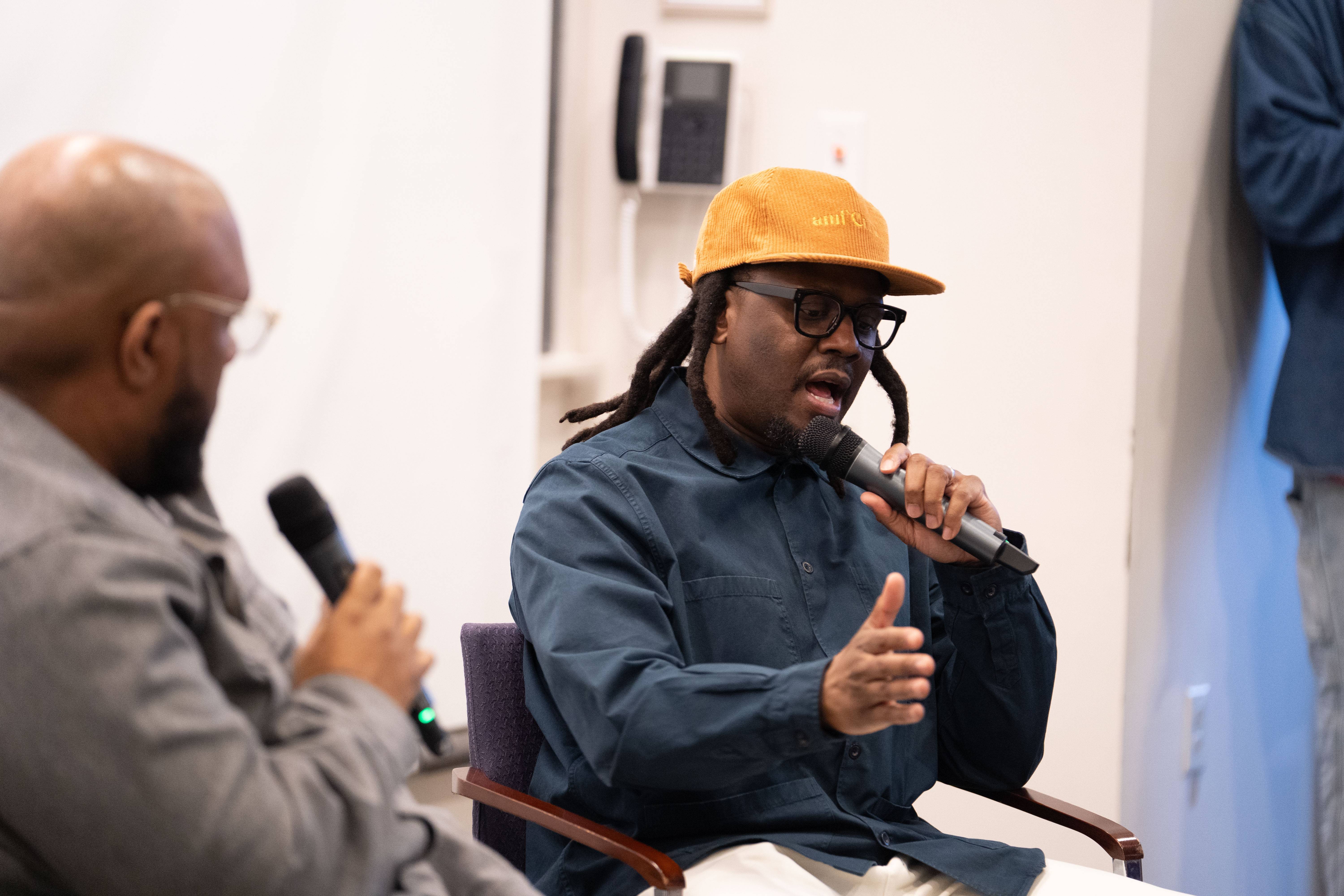 A speaker in an orange cap and glasses gestures while speaking into a microphone during a campus discussion, as another speaker listens nearby.