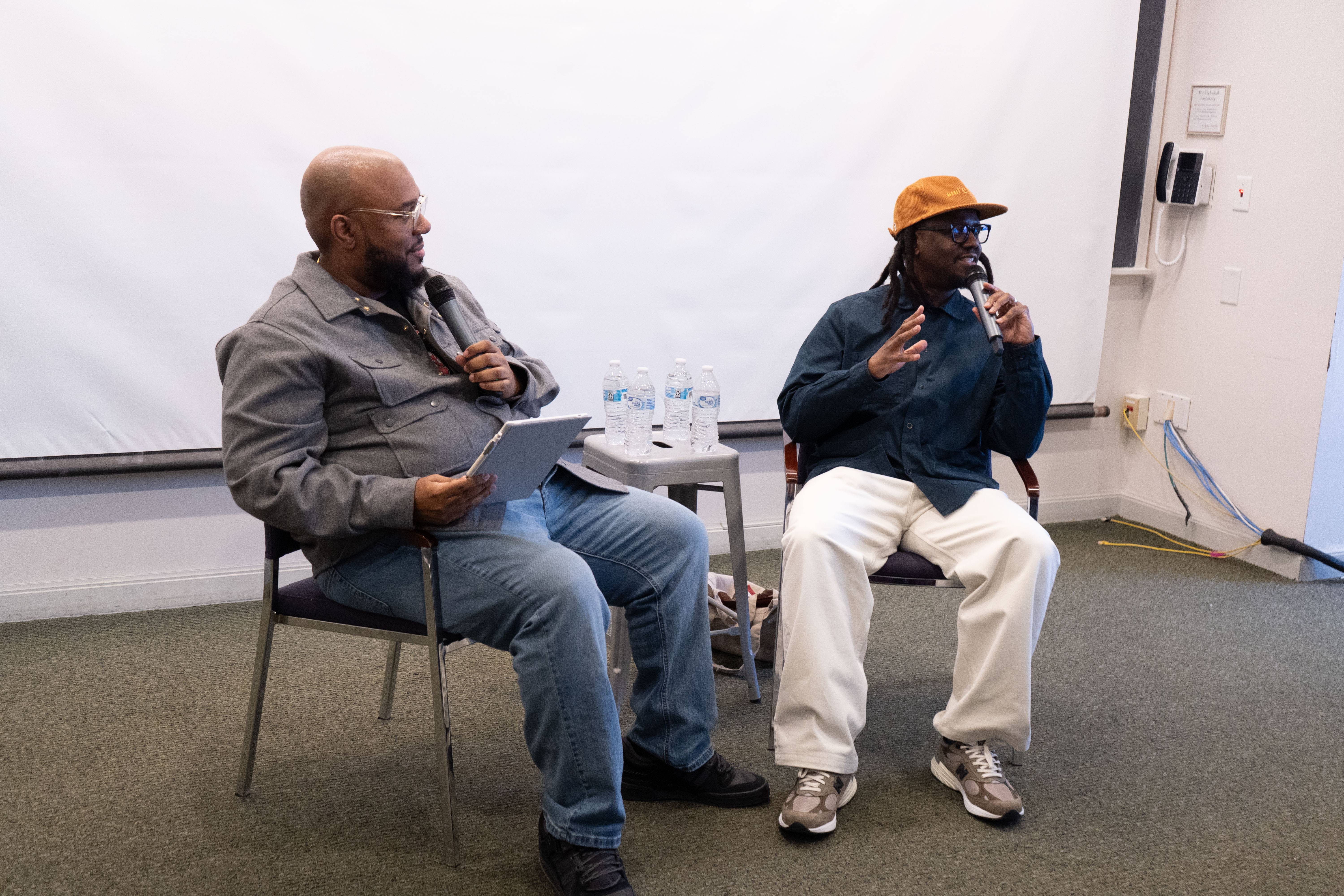 Two speakers sit at the front of a room holding microphones during a discussion, with a small table of water bottles between them and a projection screen behind them.