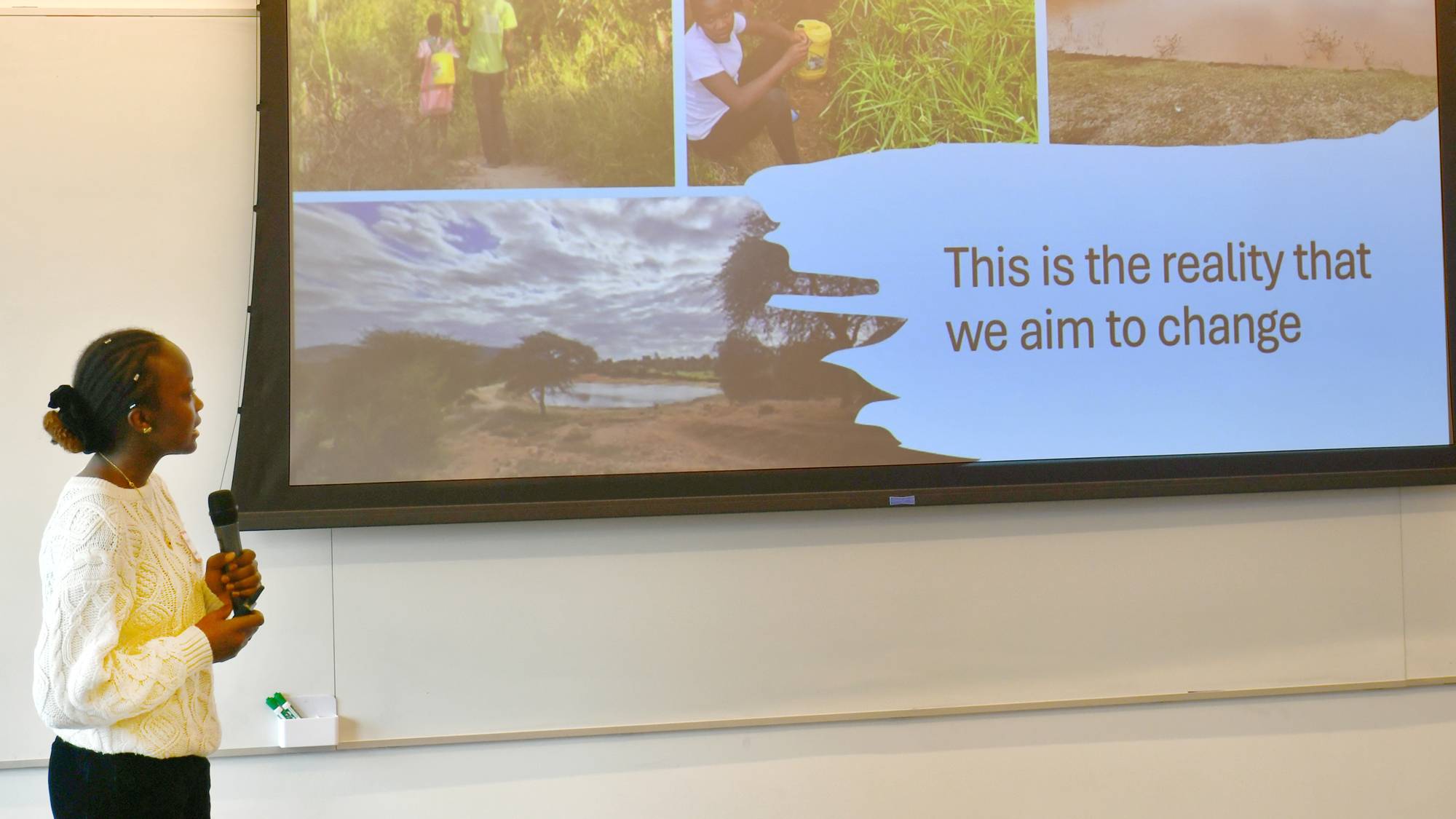 A young woman stands at the front of a room, holding a microphone and gesturing toward a large presentation screen that displays images of rural landscapes and the text, "This is the reality that we aim to change."
