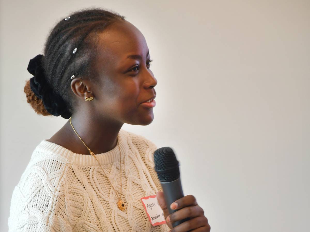A young woman with braided hair and a white cable-knit sweater speaks into a handheld microphone.