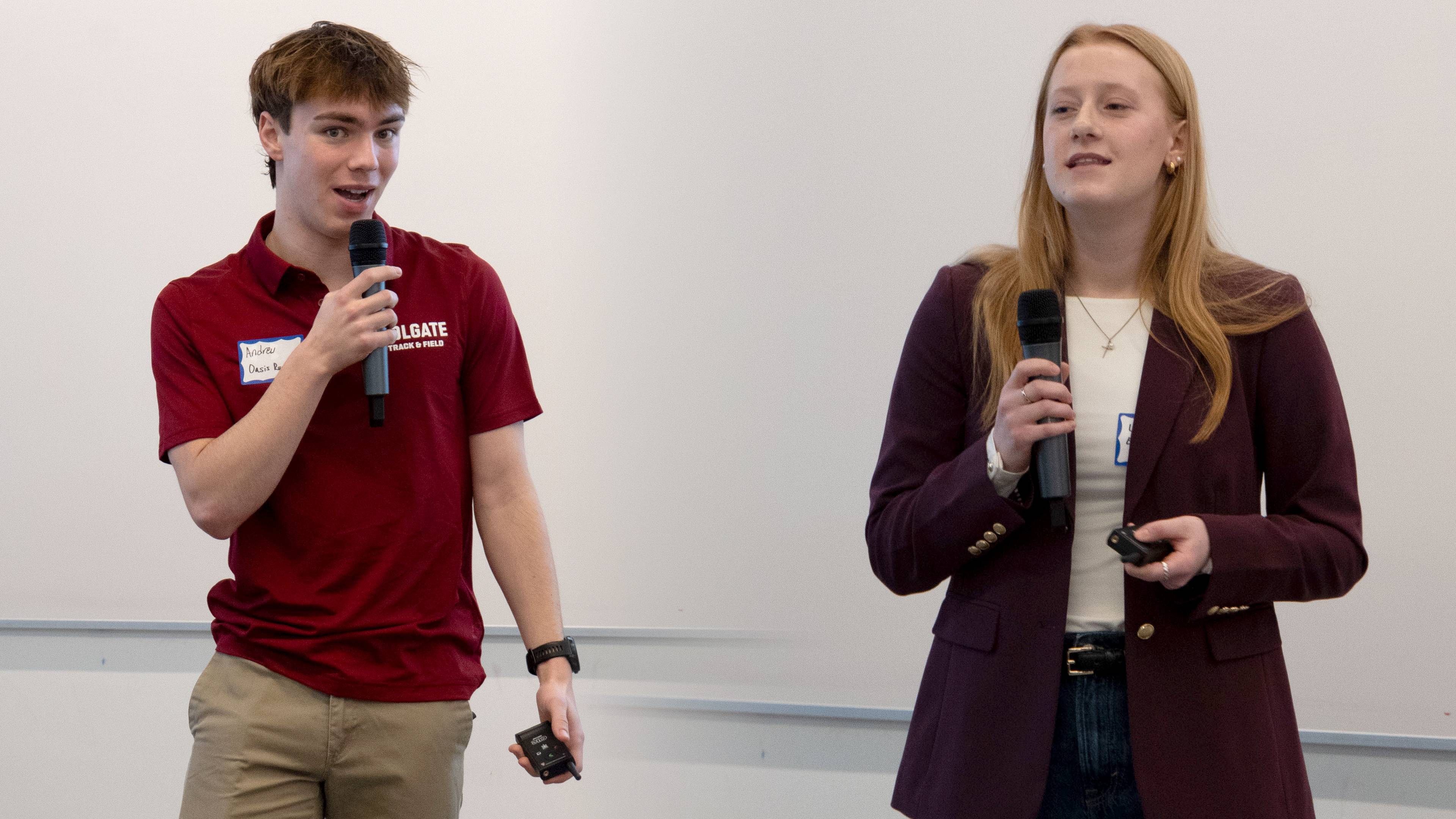 Two student entrepreneurs stand in front of a whiteboard holding microphones, with one in a maroon polo speaking while the other in a purple blazer presents beside them.