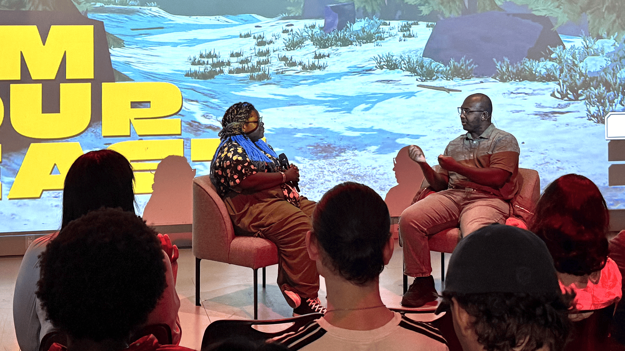 Two speakers sit in armchairs onstage in front of an audience, with a video game scene projected on a large screen behind them.