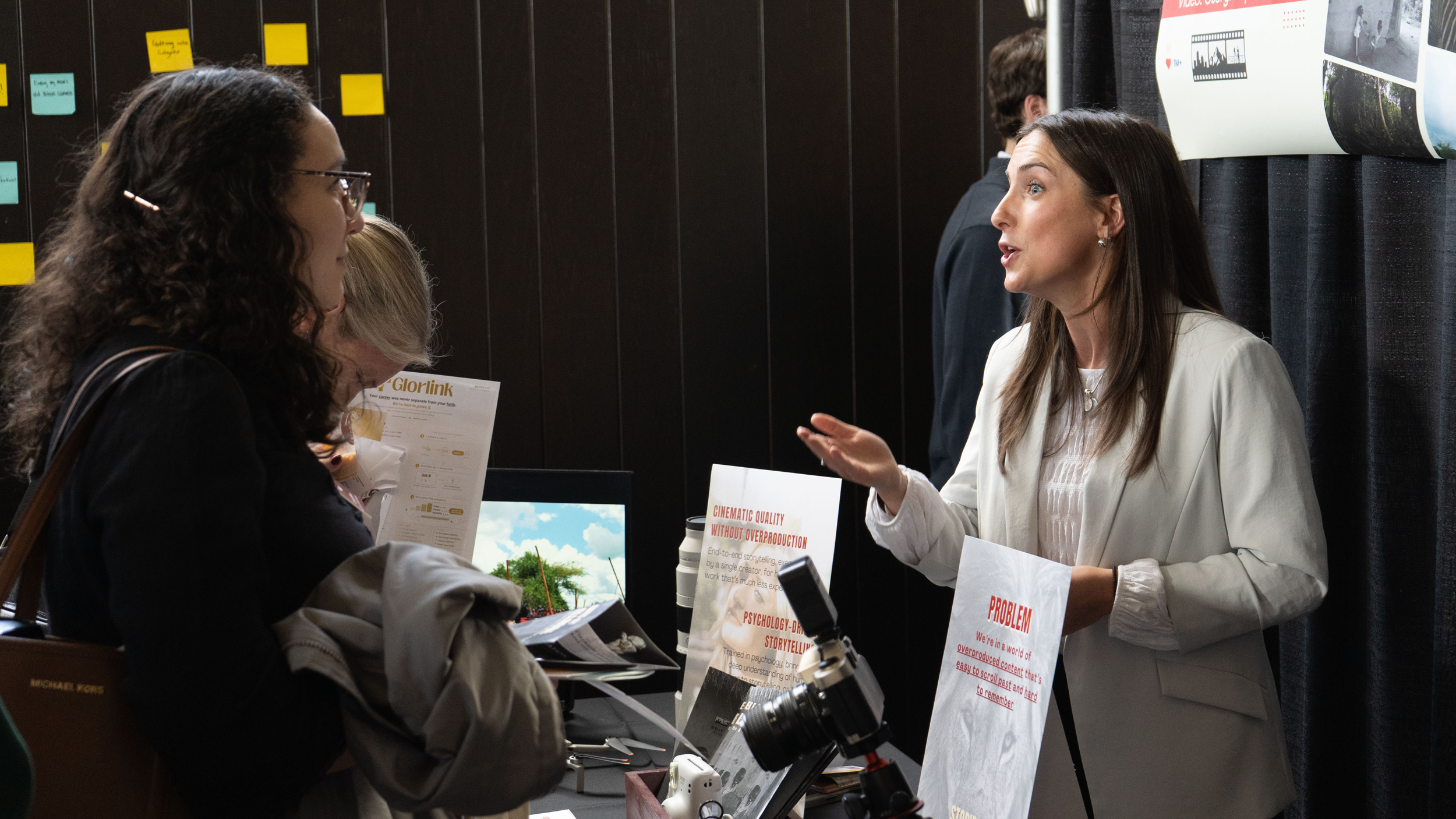 A woman wearing a white blazer gestures expressively as she speaks to an attentive listener across a display table. The booth table between them holds promotional signboards, printed flyers, and various pieces of camera equipment.