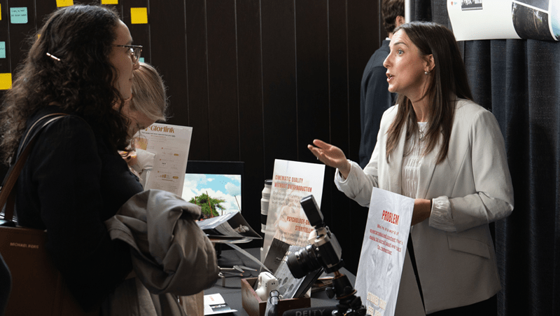 A woman wearing a white blazer gestures expressively as she speaks to an attentive listener across a display table. The booth table between them holds promotional signboards, printed flyers, and various pieces of camera equipment.