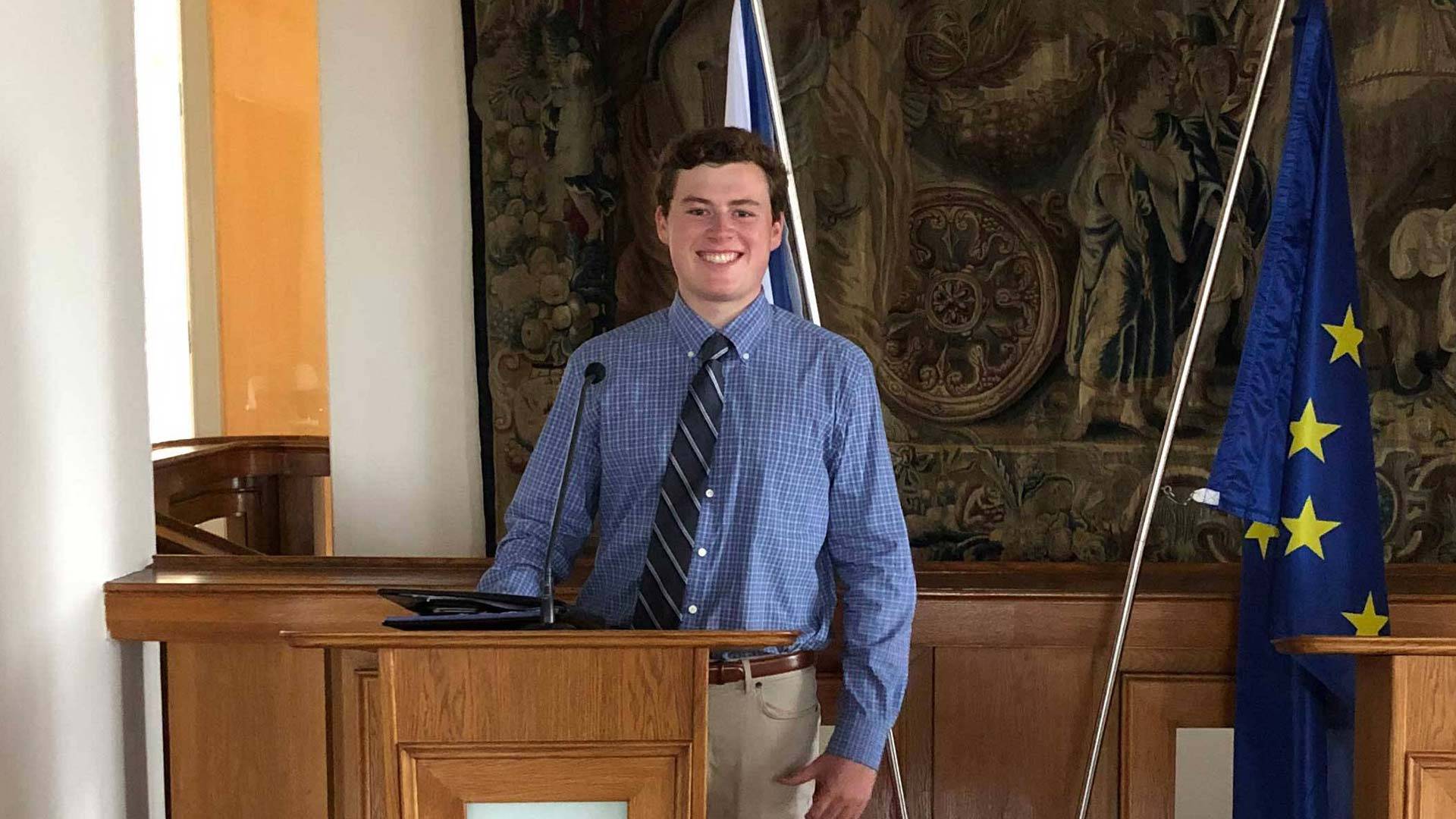 Chris Burke ’21 at a podium with the flag of the European Union behind him