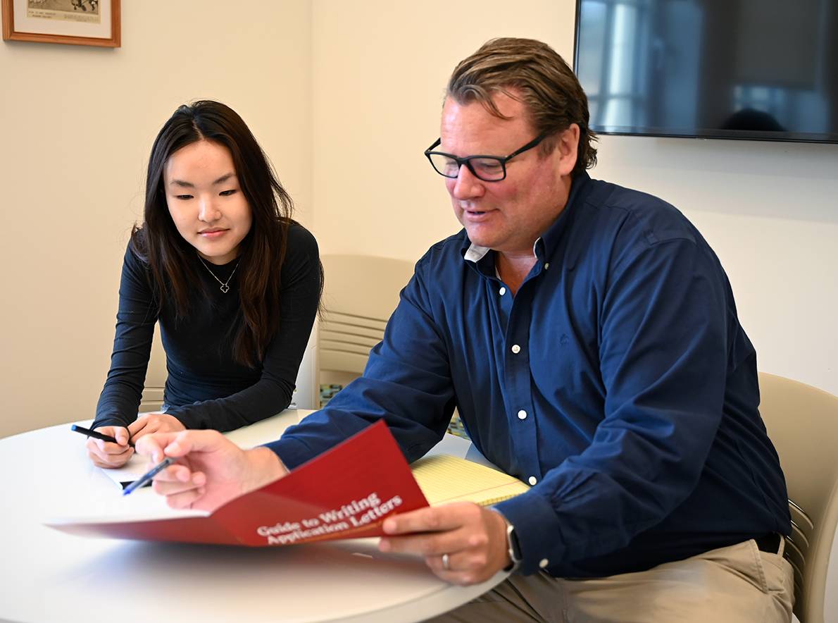 A career adviser sits at a desk with a student, going over some documents in a sunny room.