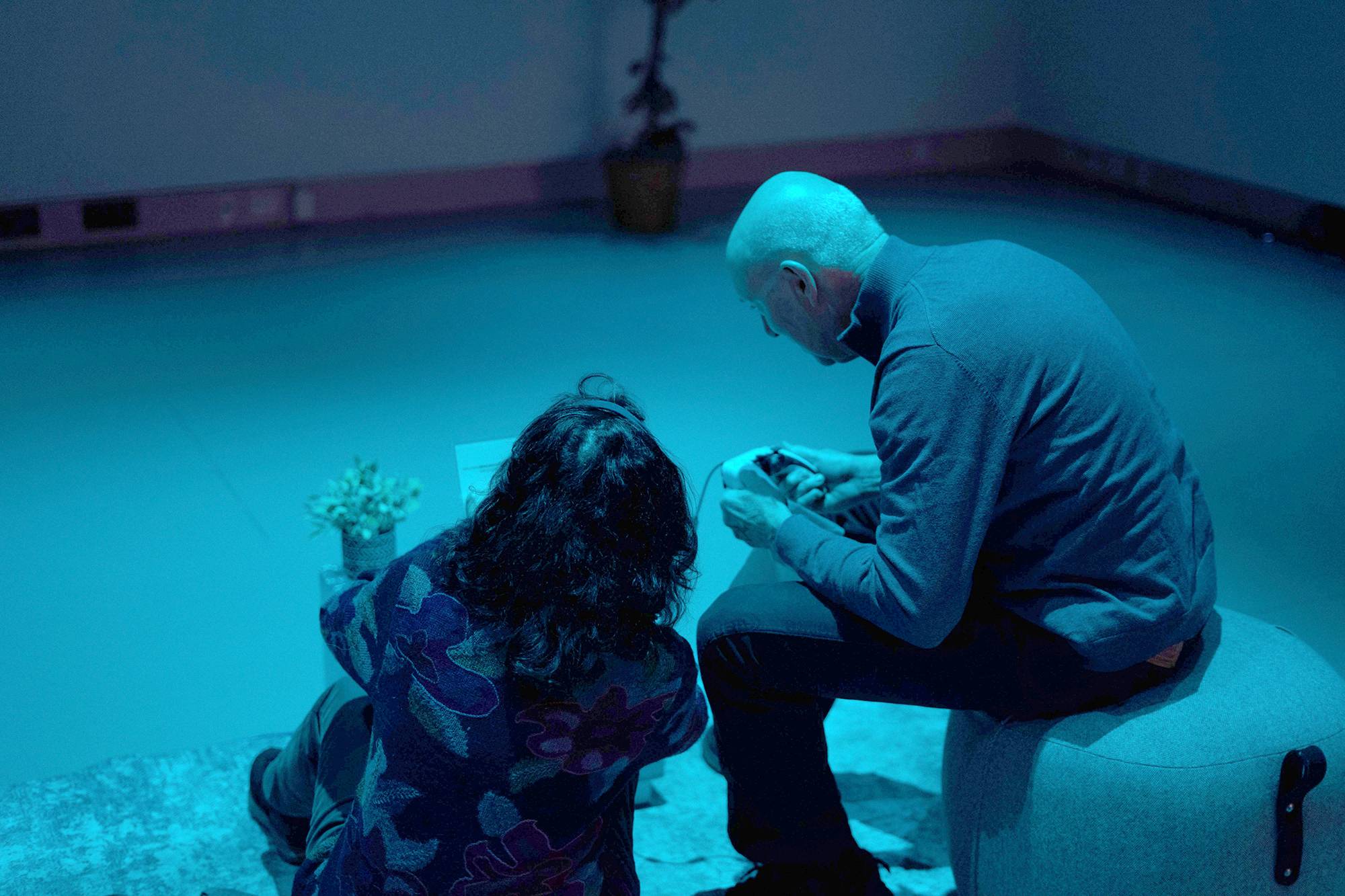 Two people sit on the floor in a dim, blue-lit room, with one person on an ottoman holding and adjusting a game controller. A small potted plant and a rug are visible nearby. 