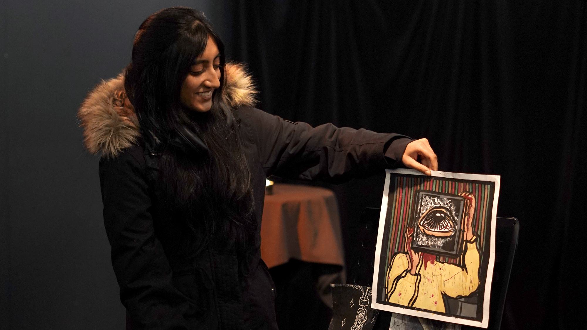 An artist in a winter coat stands with her arm extended holding a creation of hers. The room is dark behind her.