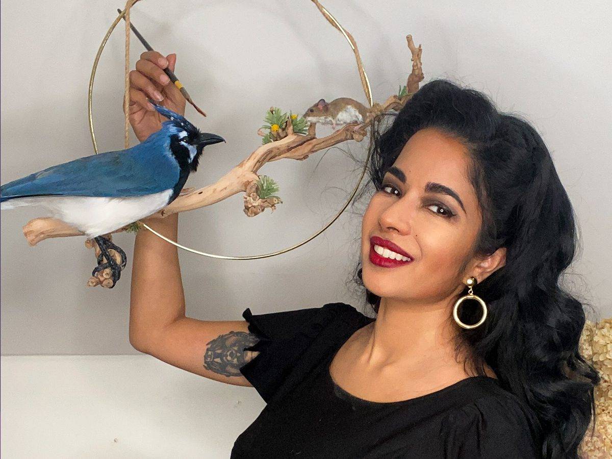 Taxidermist Divya Anantharaman smiling next to a taxidermy display featuring a bright blue bird and a small mouse perched on a branch within a circular brass ring.
