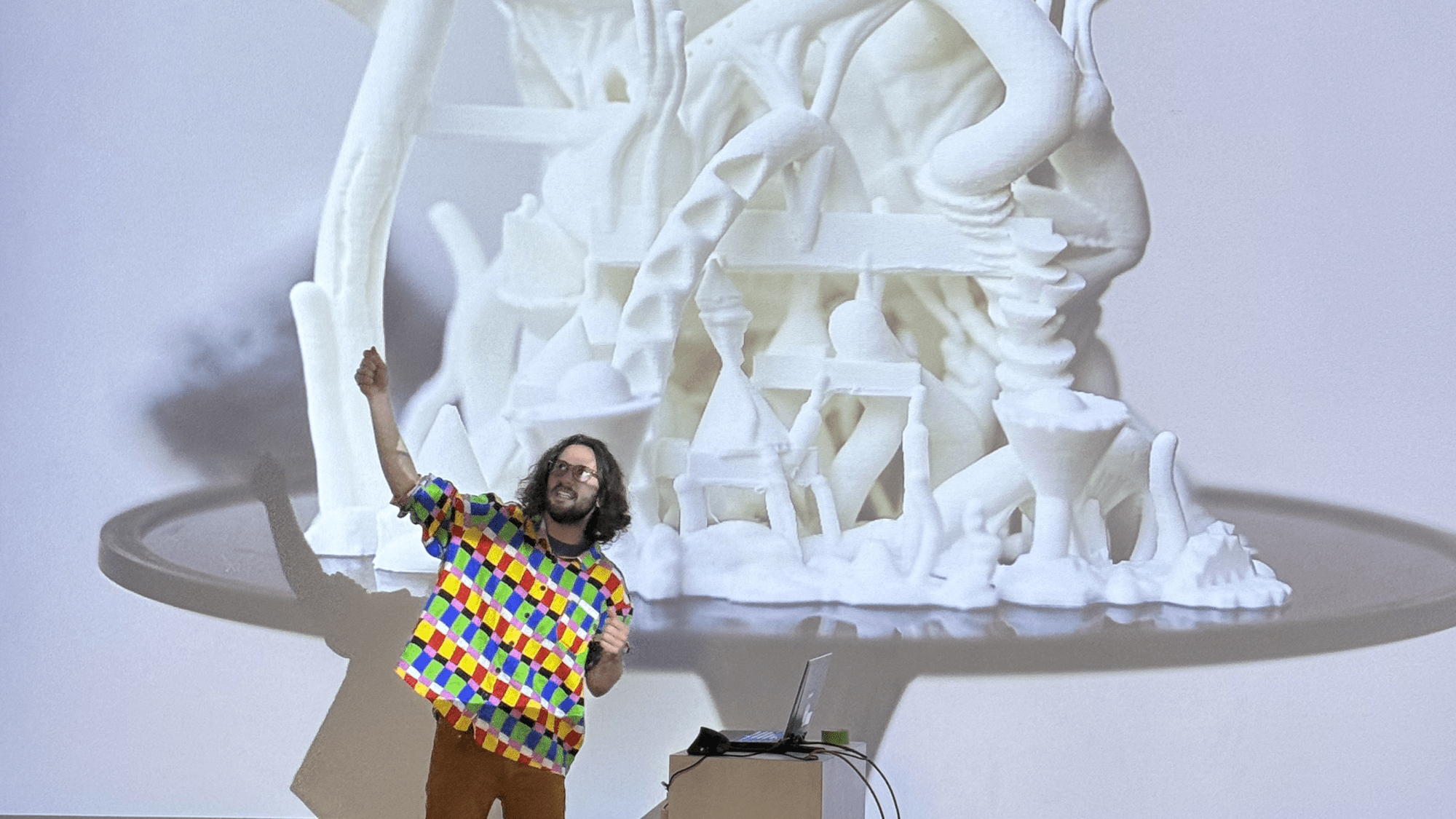 Shawn Chiki, in a brightly multicolored shirt, stands in front of a projected image of a digitally-sculpted architecture cast in white.
